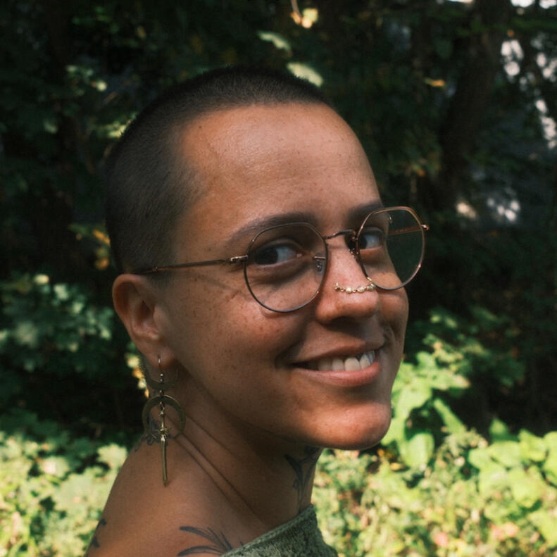 Nellie Grant, LMT A photo of Nellie Grant, the owner and sole practitioner at Cloudbelly Bodywork LLC. The photo shows their smiling face turned toward the camera with warmth and comfort in their brown eyes. They are wearing wire-framed glasses, dangly earrings, and a their