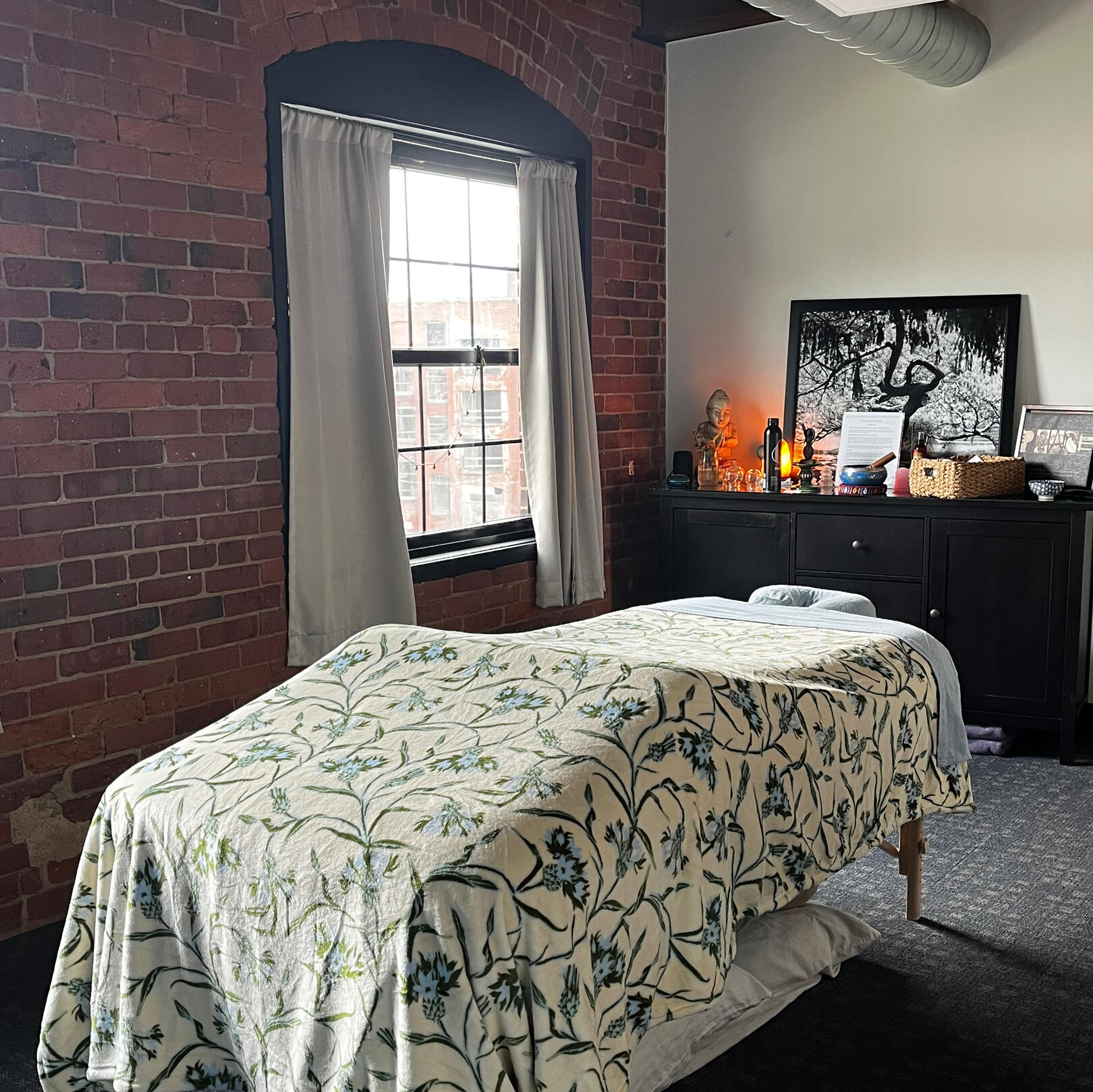 A photo of the Cloudbelly Bodywork LLC studio. The photo shows a massage table at the center. It's covered with a cream-colored blanket that has green vines and flowers with light and dark blue accents.
